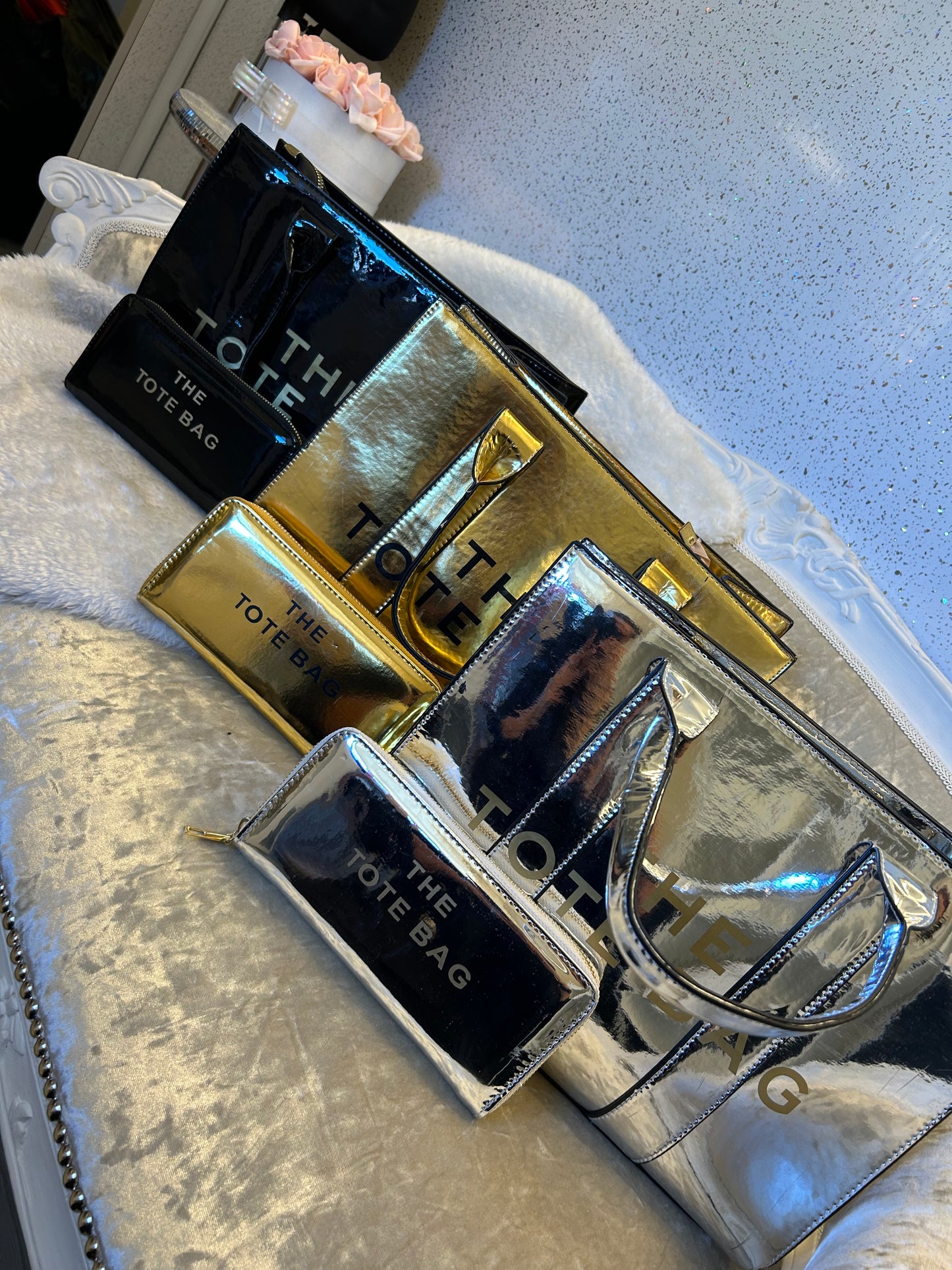 Collection of 'The Tote Bag' products including a black bag, gold clutch, and silver handbag on a textured surface.