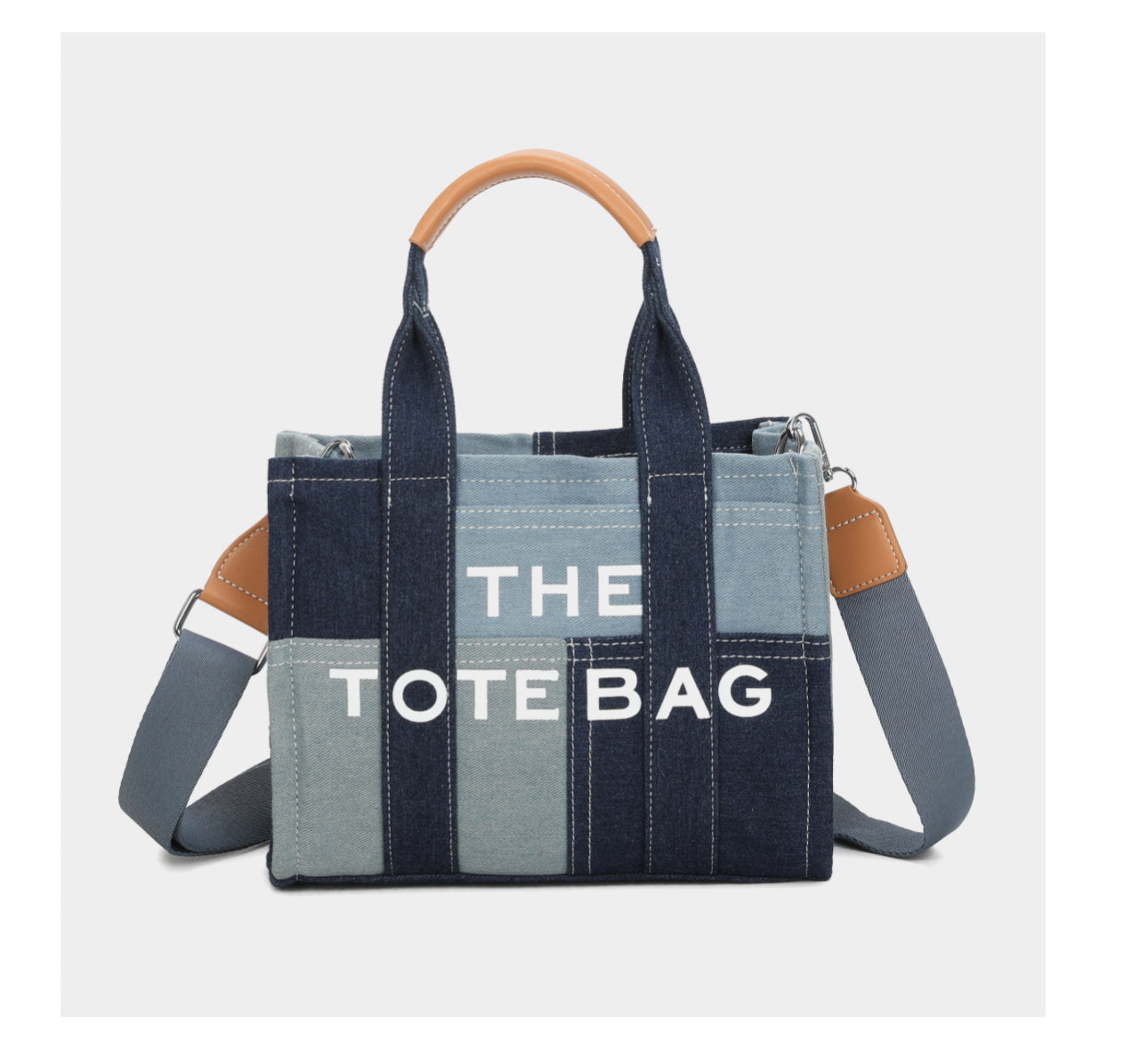 A blue denim patchwork tote handbag with 'THE TOTE BAG' text on one of the patches, featuring a brown handle and an adjustable crossbody strap.