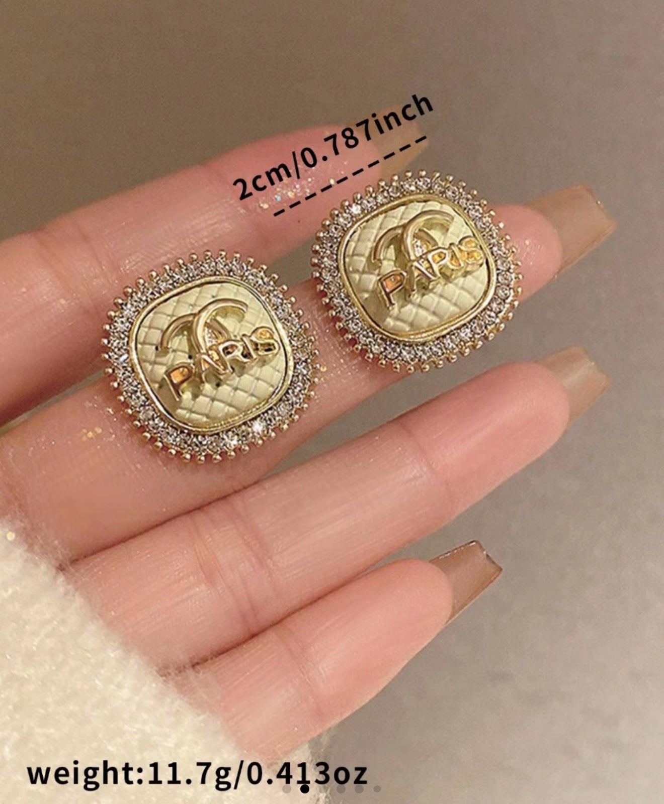 Gold and diamond earrings held on a hand with measurement label