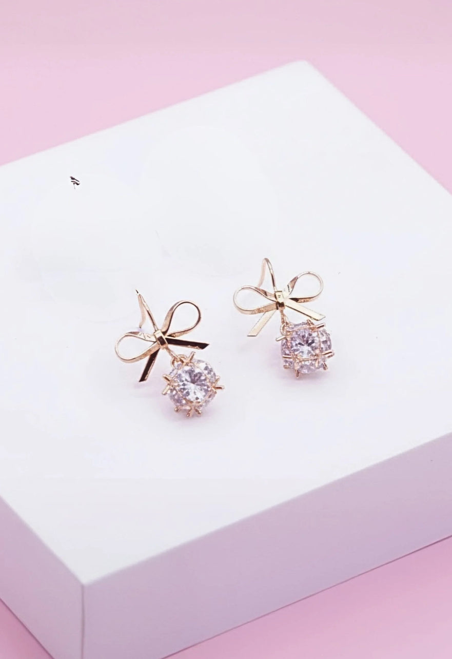 Rose gold earrings with bow designs on a white box with pink background