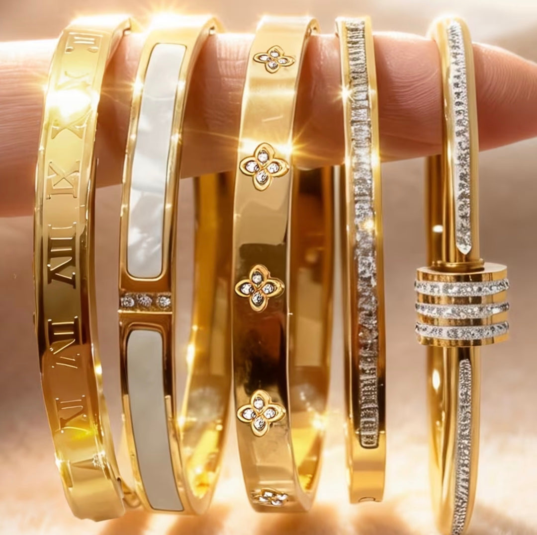 Five-piece gold-tone bangle bracelet set with crystal detailing, Roman numeral accents and clover design stacked on wrist.