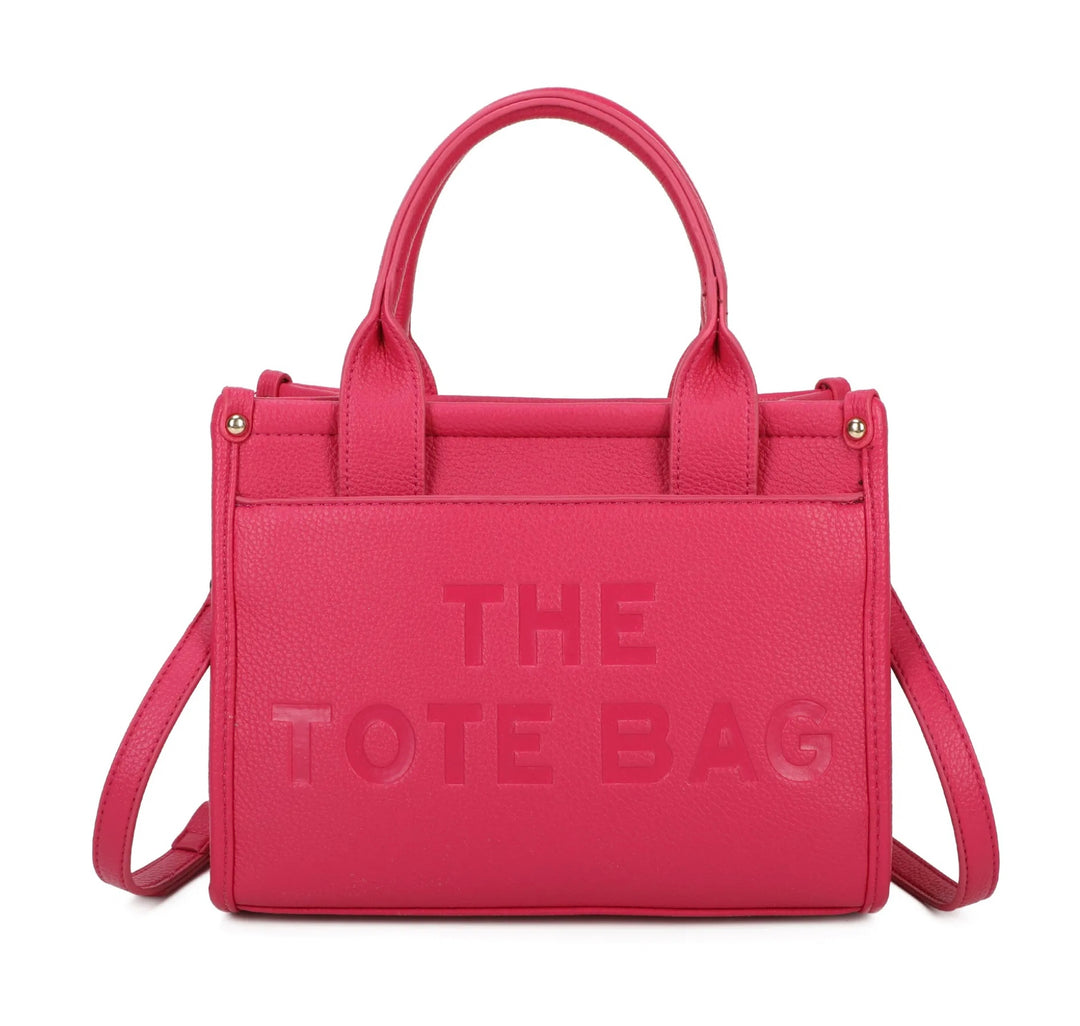 A pink tote bag with the text 'THE TOTE BAG' embossed on the front, featuring a front pocket, double grab straps, and gold tone metal hardware.