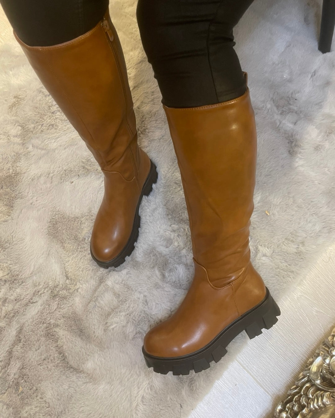 Tan knee high boots with black sole worn on feet, stylish women’s autumn footwear