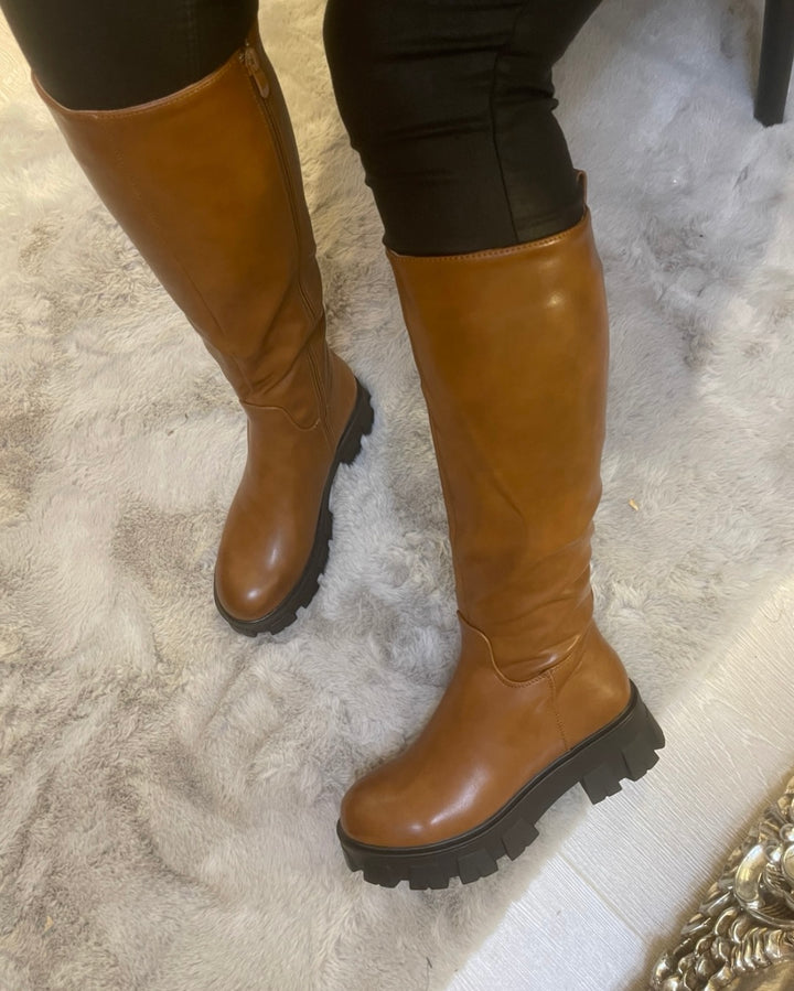 Tan knee high boots with black sole worn on feet, stylish women’s autumn footwear