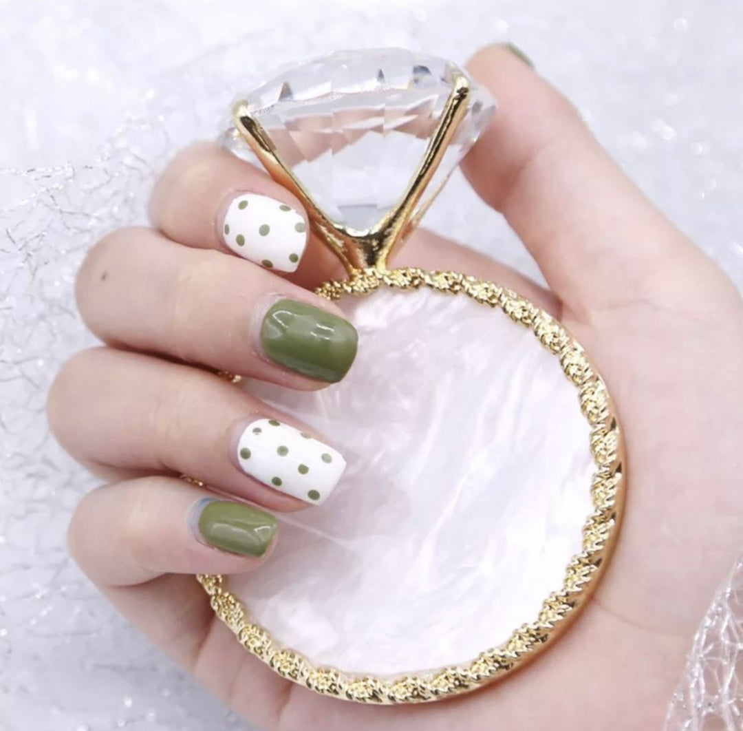 A hand holding a gold-colored nail art palette with various colorful nail polish shades and a single large clear crystal head.