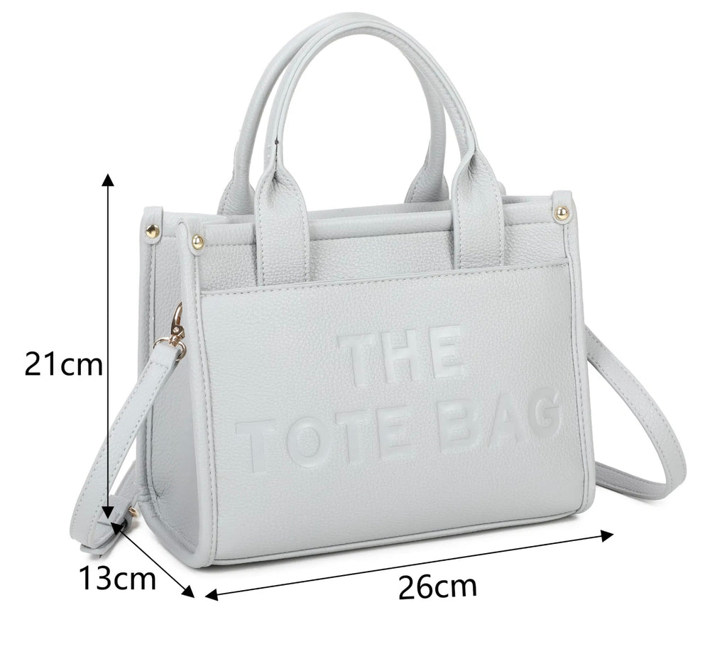 A grey tote bag with the text 'THE TOTE BAG' embossed on the front, featuring a front pocket, double grab straps, and gold tone metal hardware.