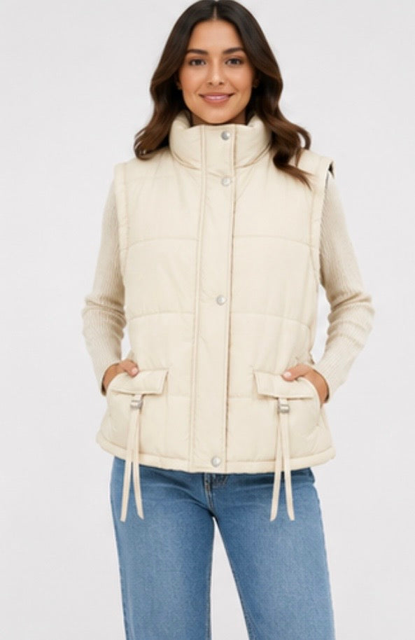 Cream sleeveless padded gilet with high neck, front popper fastening, quilted design, adjustable buckle pocket details and elasticated waist