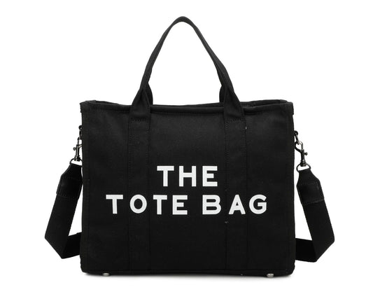 A black canvas tote bag with 'THE TOTE BAG' printed in white text. It features double grab straps, a detachable and adjustable strap, and silver tone metal hardware.