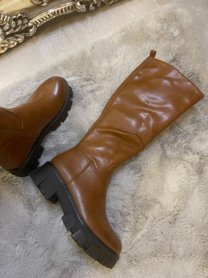 Tan knee high boots with black sole worn on feet, stylish women’s autumn footwear
