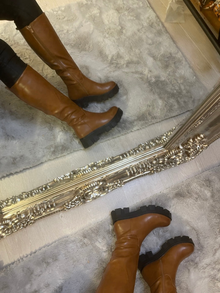 Tan knee high boots with black sole worn on feet, stylish women’s autumn footwear