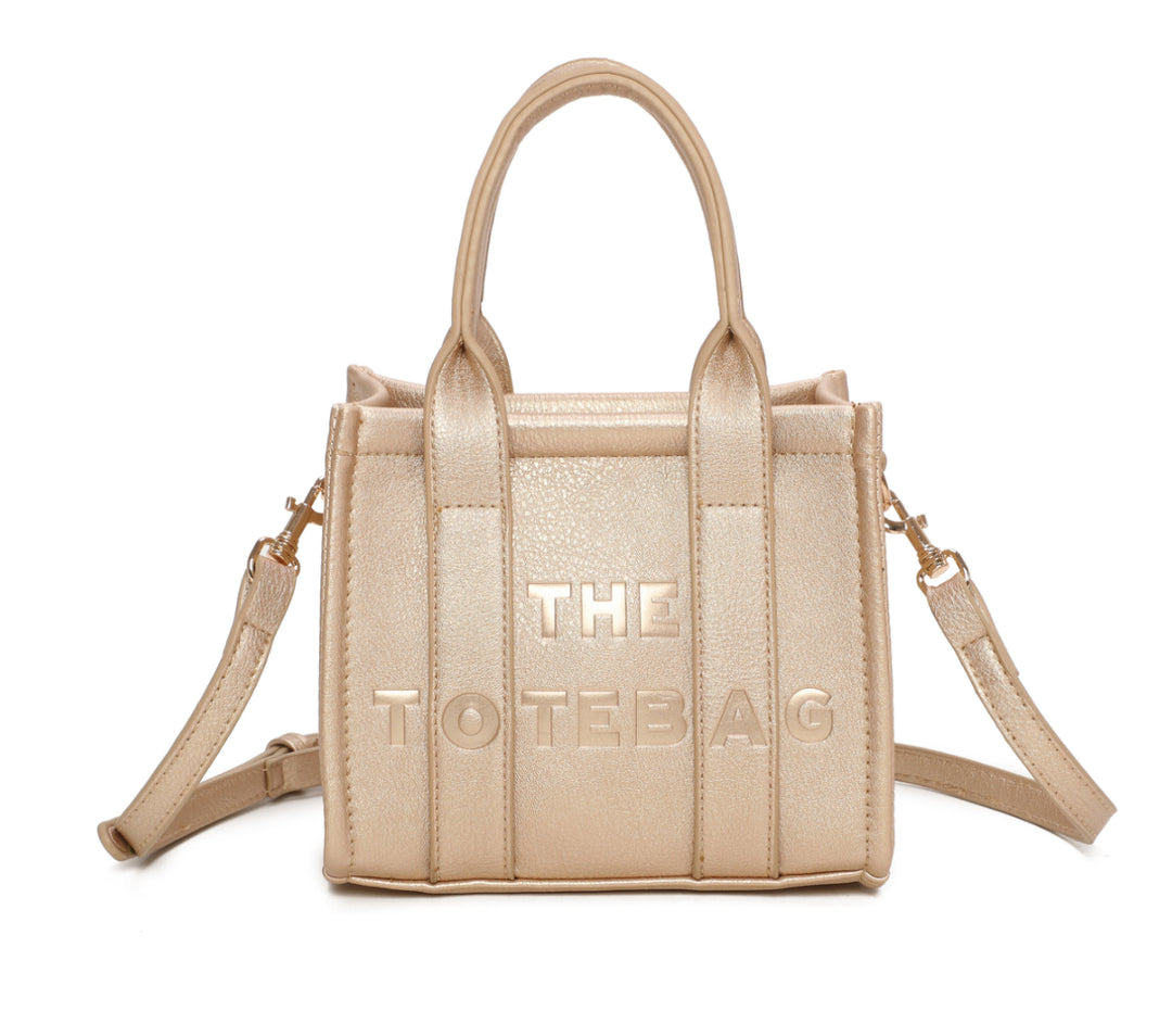 A gold mini tote bag with 'THE TOTE BAG' embossed on the front, featuring double grab handles and a detachable strap.