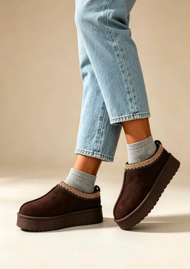 Dark brown suede platform slipper boots with a decorative beige braided trim, shown on a model wearing light blue jeans and gray socks against a warm beige background.