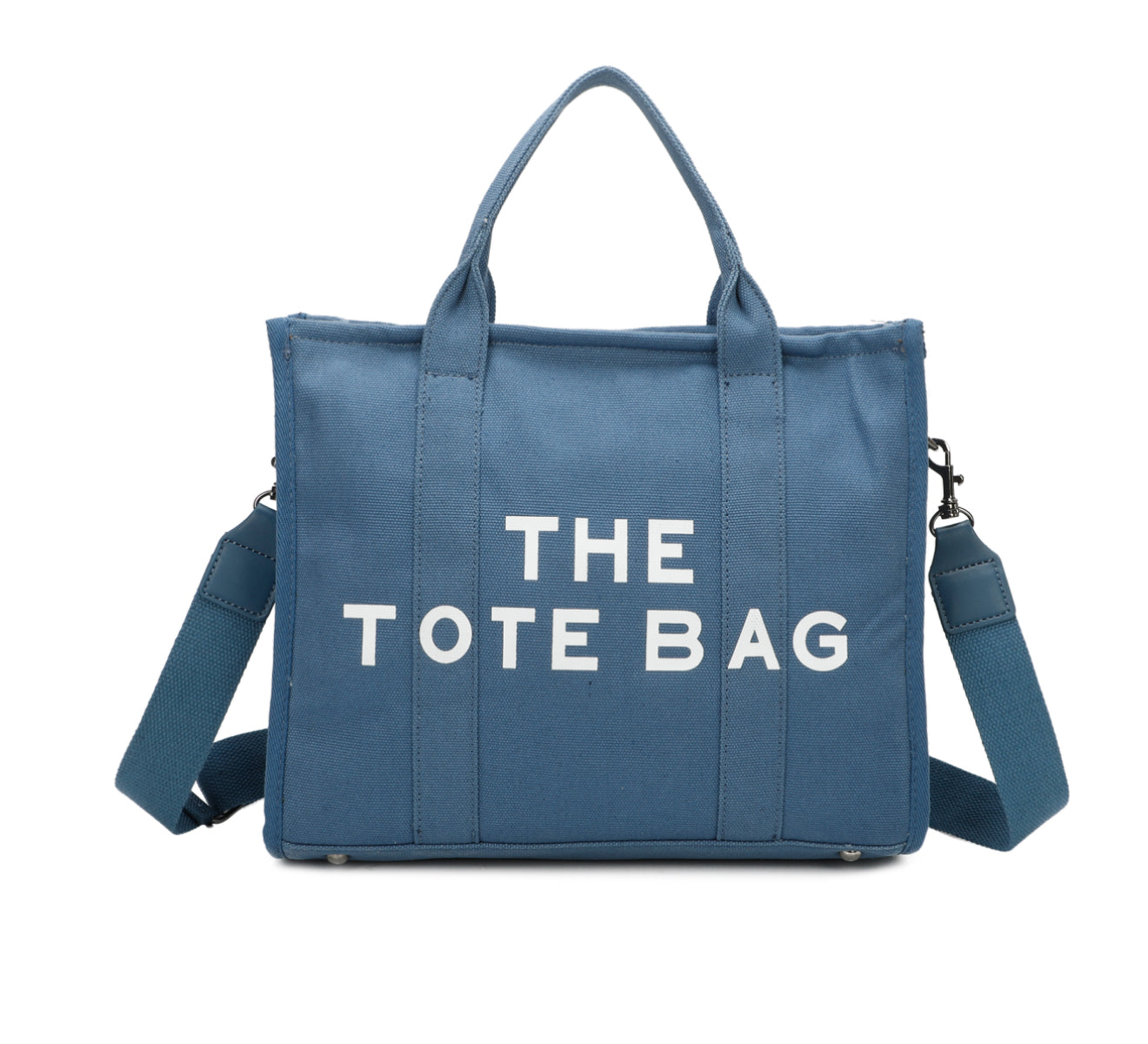 A blue canvas tote bag with 'THE TOTE BAG' printed in white text. It features double grab straps, a detachable and adjustable strap, and silver tone metal hardware.