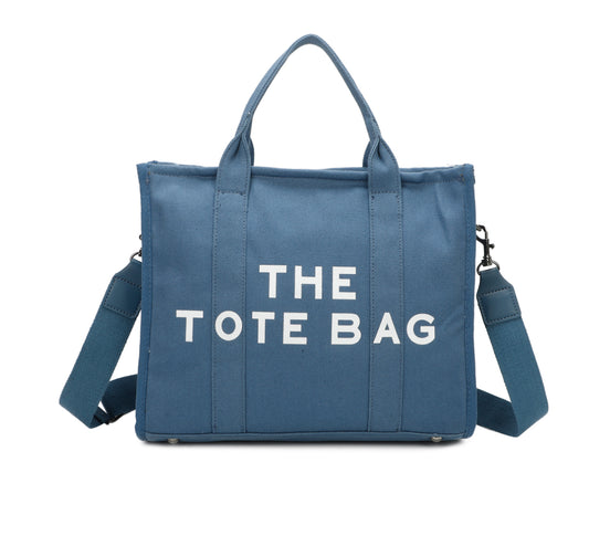 A blue canvas tote bag with 'THE TOTE BAG' printed in white text. It features double grab straps, a detachable and adjustable strap, and silver tone metal hardware.