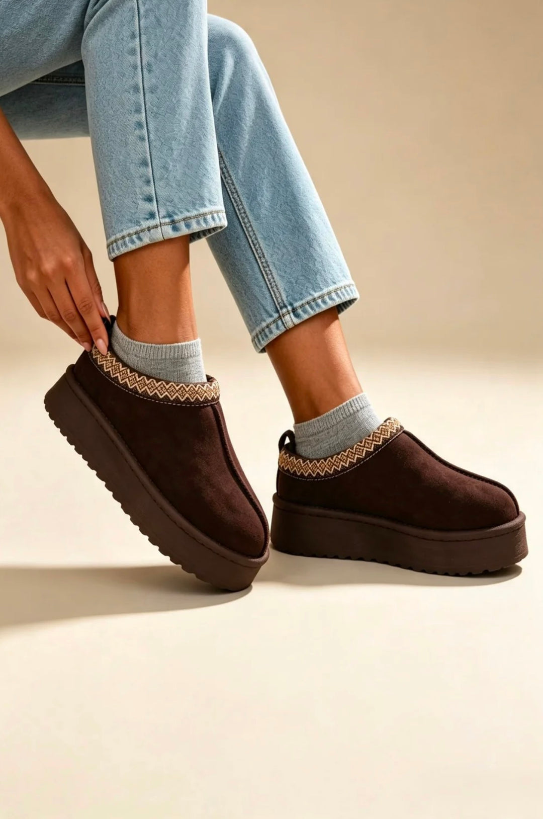 A person wearing dark brown platform UGG-style slippers with a beige braided trim, paired with light blue jeans and gray socks, shown against a warm beige studio background.