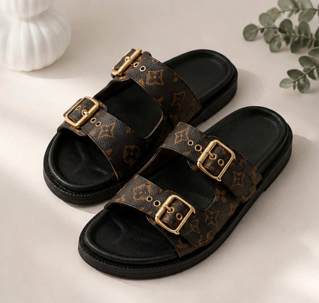 A pair of black double-strap slide sandals with brown monogram-patterned straps and gold-tone buckles, styled on a light beige surface with a white decorative object and green leaves in the background.