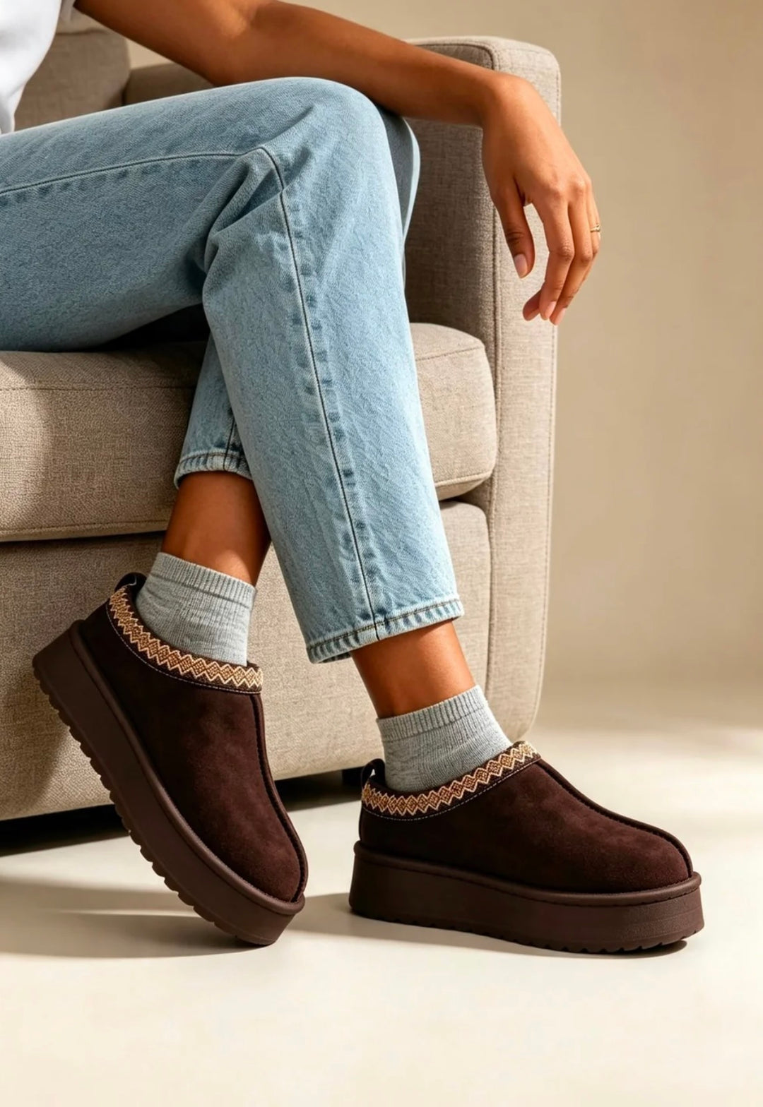 A person seated on a light beige armchair wearing dark brown platform UGG-style slipper boots with decorative beige trim and light gray socks, against a neutral indoor background.