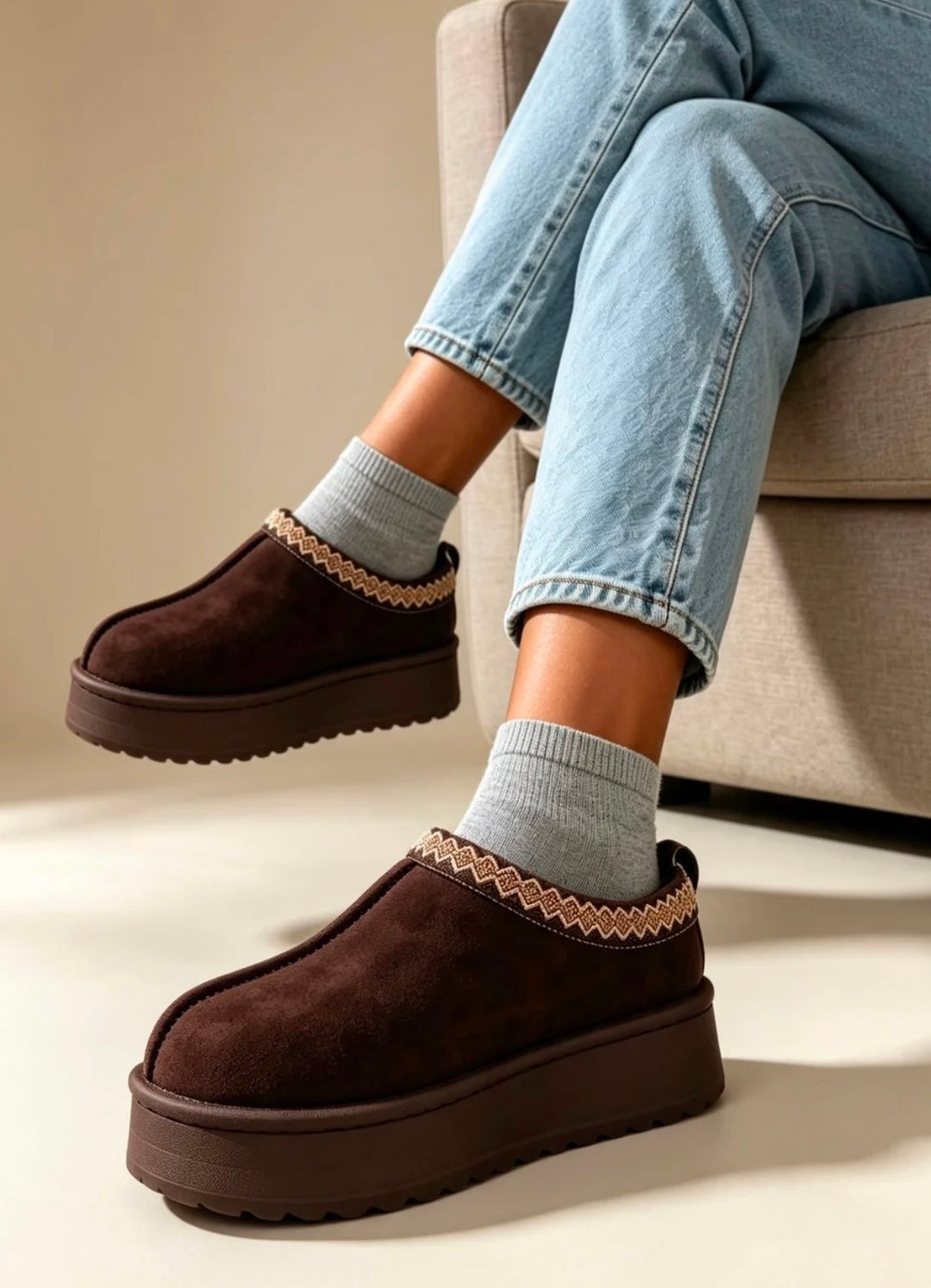 A person seated on a light beige sofa wearing dark brown suede slip-on platform slippers with a decorative braided trim and light gray socks, paired with light blue jeans.