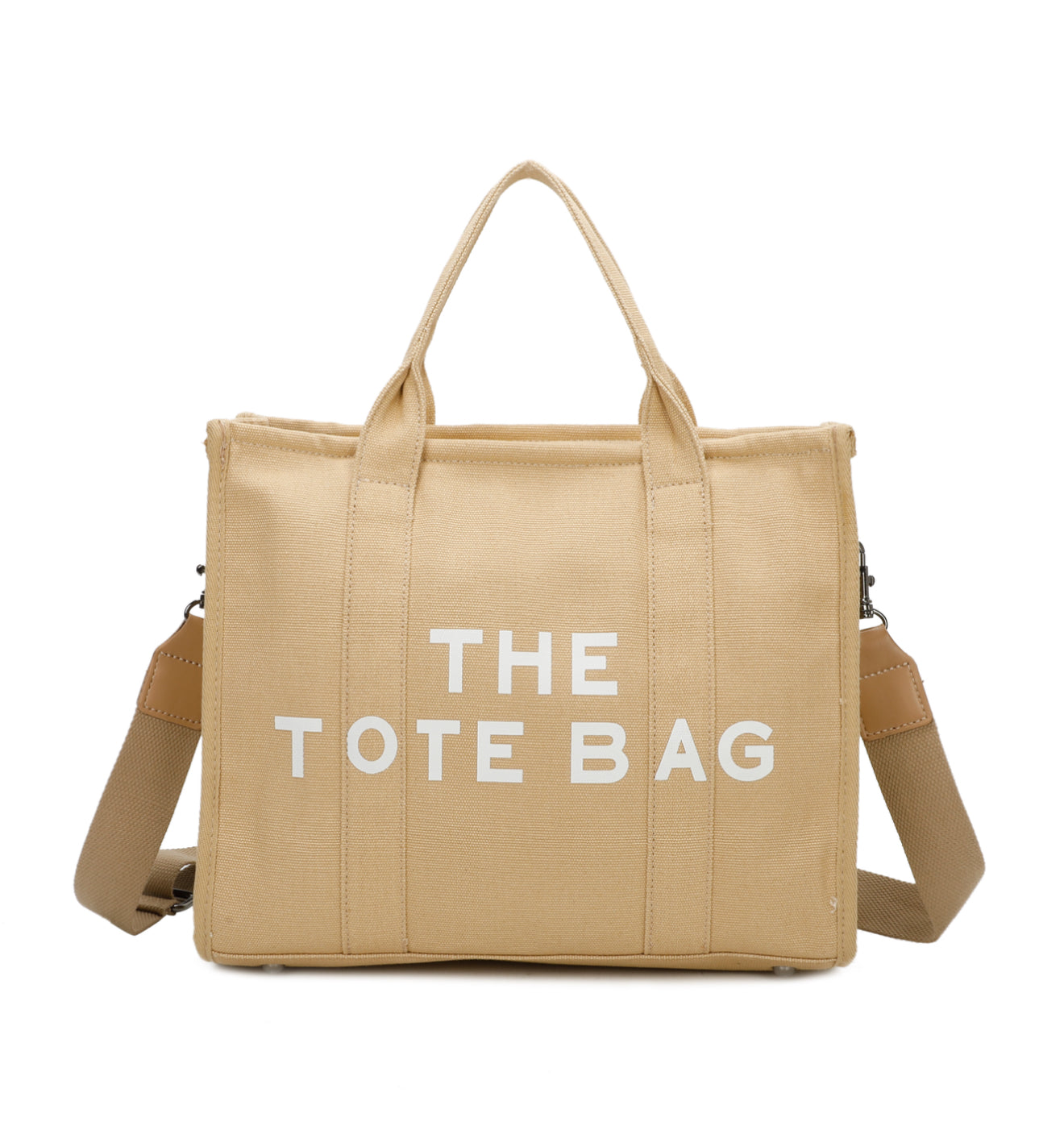 A beige canvas tote bag with 'THE TOTE BAG' printed in white text. It features double grab straps, a detachable and adjustable strap, and silver tone metal hardware
