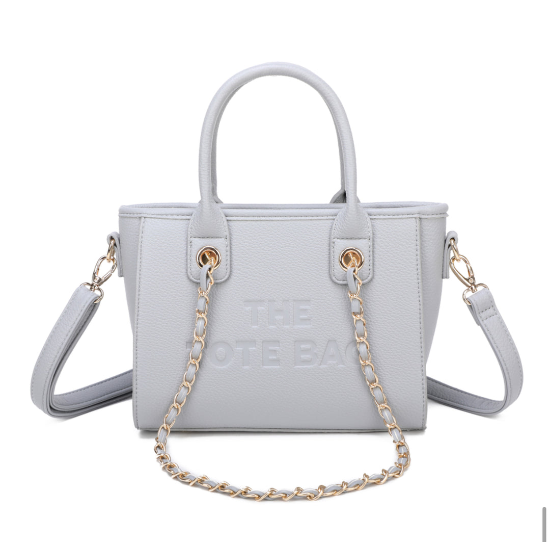 A grey tote bag with a chain handle, featuring a front zipper pocket and gold tone metal hardware. The &