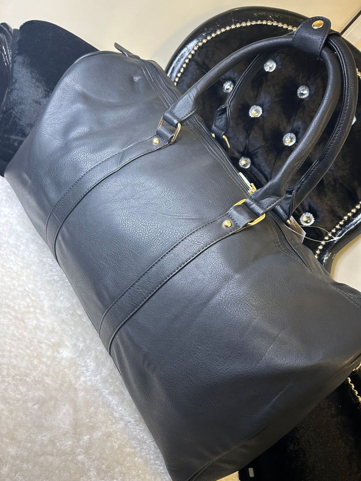 A black luggage holdall with gold detail, featuring a barrel shape, twin grab handles, and a detachable shoulder strap.