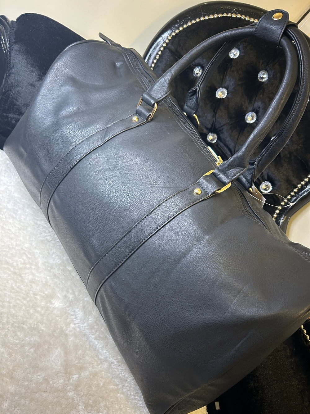 A black luggage holdall with gold detail, featuring a barrel shape, twin grab handles, and a detachable shoulder strap.