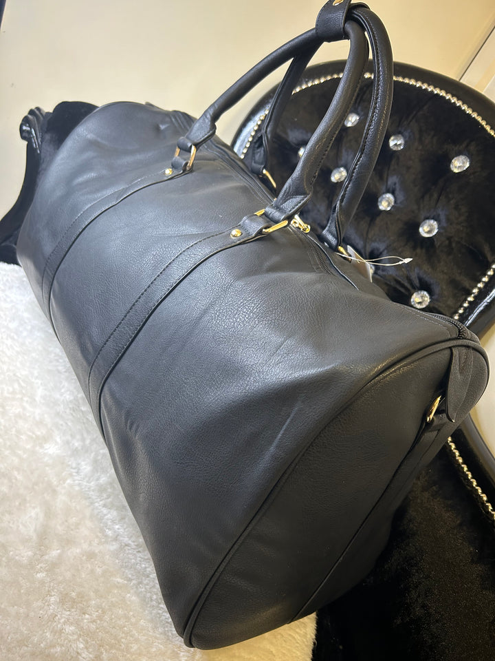 A black luggage holdall with gold detail, featuring a barrel shape, twin grab handles, and a detachable shoulder strap.