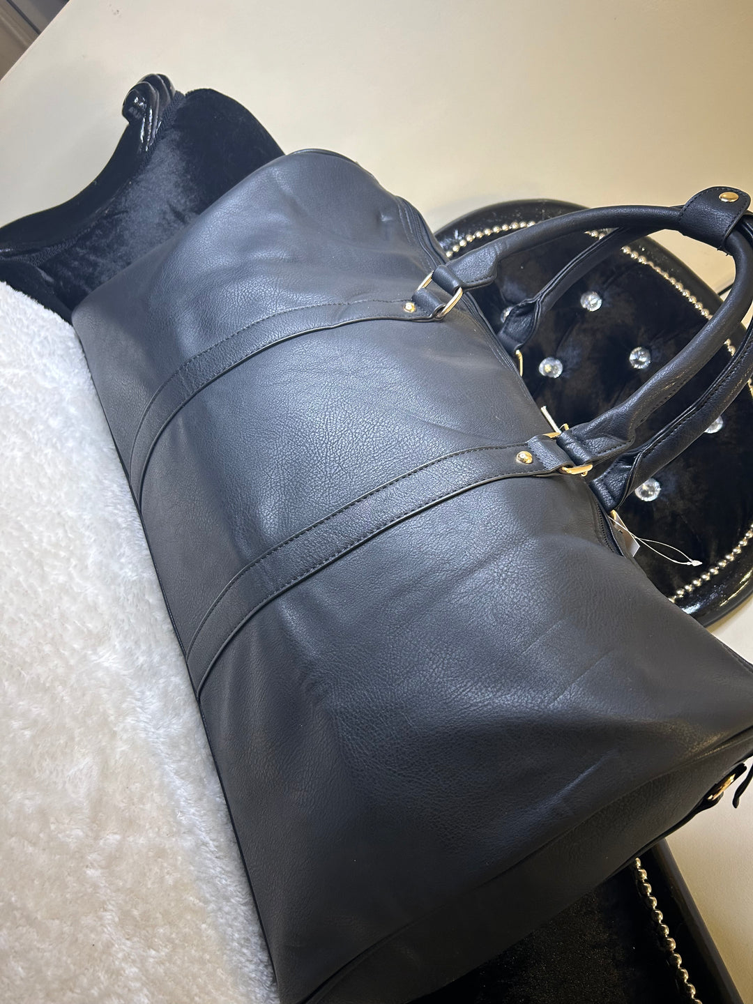A black luggage holdall with gold detail, featuring a barrel shape, twin grab handles, and a detachable shoulder strap.